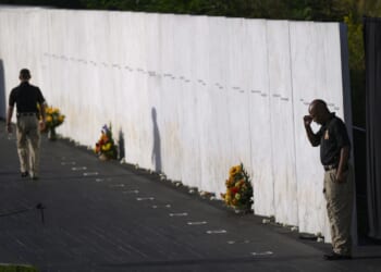 Two security guards stand by a white marble memorial wall with engraved names and flowers placed at its base on a sunny day.
