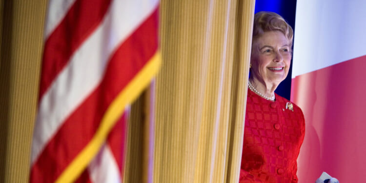 A woman in a red dress stands behind a curtain, smiling. An out-of-focus American flag is visible in the foreground.
