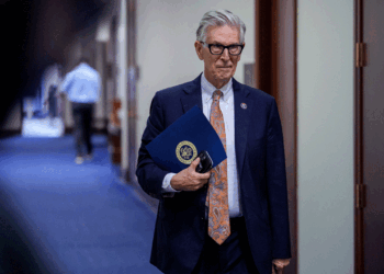 An older man in a suit and glasses holds a blue folder and sunglasses, walking down a hallway with blue carpet and wooden doors—perhaps on his way to discuss the latest developments in AI.