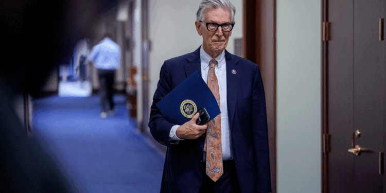 An older man in a suit and glasses holds a blue folder and sunglasses, walking down a hallway with blue carpet and wooden doors—perhaps on his way to discuss the latest developments in AI.