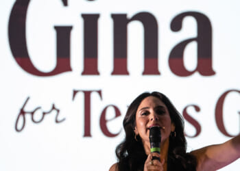 A woman with dark hair speaks into a microphone, raising one finger, in front of a large screen displaying partially visible text that reads Gina and for Texas..