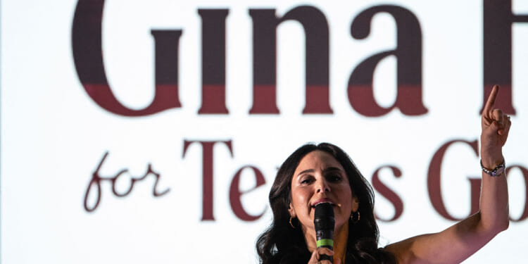 A woman with dark hair speaks into a microphone, raising one finger, in front of a large screen displaying partially visible text that reads Gina and for Texas..