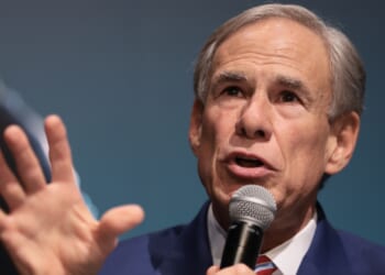 Gov. Greg Abbott in a suit speaks into a microphone, gesturing with his hand while addressing an audience, against a blue background.