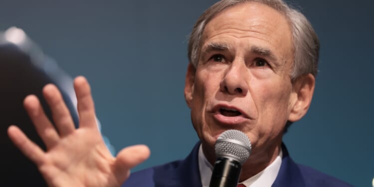 Gov. Greg Abbott in a suit speaks into a microphone, gesturing with his hand while addressing an audience, against a blue background.
