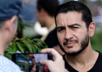 A man with a trimmed beard stands outdoors, looking slightly serious as another person in a cap takes his photo or video with a smartphone. Greenery is visible in the background.