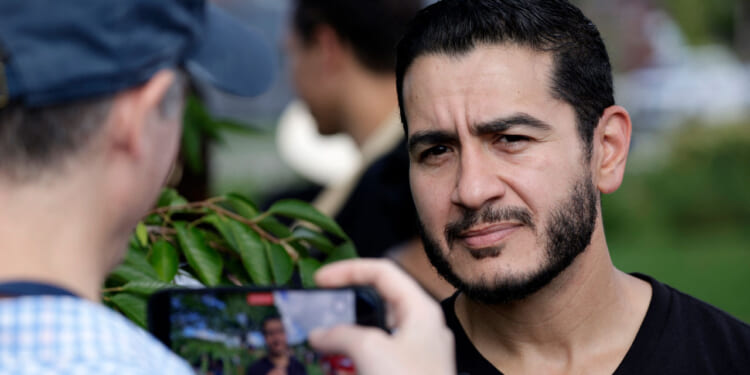 A man with a trimmed beard stands outdoors, looking slightly serious as another person in a cap takes his photo or video with a smartphone. Greenery is visible in the background.