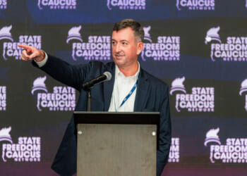 A man in a suit stands at a podium, pointing to his right, with a microphone in front of him and a backdrop reading State Freedom Caucus Network..