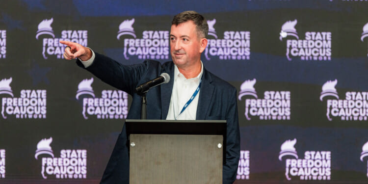 A man in a suit stands at a podium, pointing to his right, with a microphone in front of him and a backdrop reading State Freedom Caucus Network..