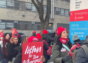 A group of people in red hats and masks hold signs saying listen to the nurses outside a hospital emergency entrance, participating in a protest or demonstration.
