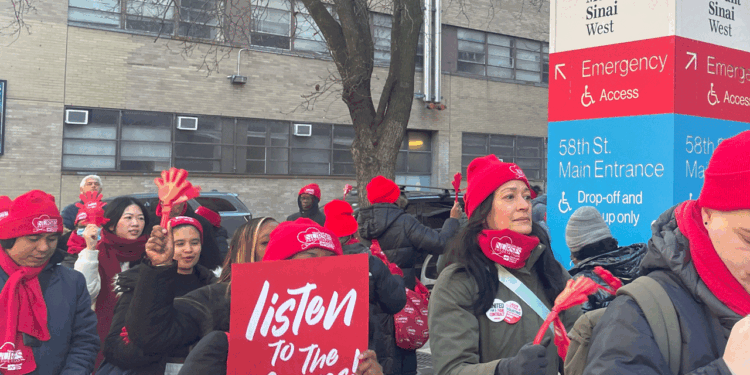 A group of people in red hats and masks hold signs saying listen to the nurses outside a hospital emergency entrance, participating in a protest or demonstration.