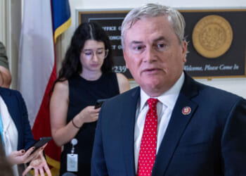 A man in a suit and red tie speaks while three women stand behind him, one holding a phone. Office plaques and flags are visible in the background.