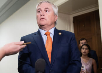 James Comer in a blue suit and orange tie stands in a hallway, surrounded by people and microphones, as reporters hold out recording devices and take notes.