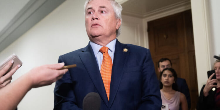 James Comer in a blue suit and orange tie stands in a hallway, surrounded by people and microphones, as reporters hold out recording devices and take notes.