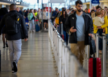 Crowds of travelers with luggage wait in line and walk through a busy airport terminal, all preparing for flying to their destinations. A person in a yellow coat holds a child on the right side of the image.