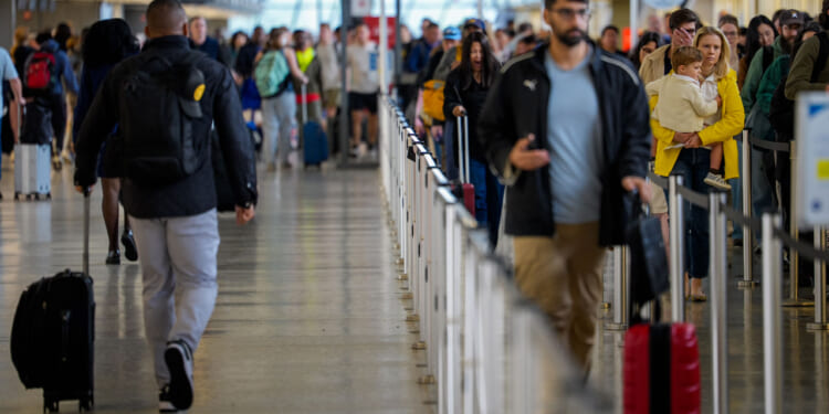 Crowds of travelers with luggage wait in line and walk through a busy airport terminal, all preparing for flying to their destinations. A person in a yellow coat holds a child on the right side of the image.