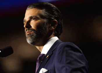 Don Trump Jr. in a suit and tie with slicked-back hair and a beard stands at a microphone, looking serious, with a dark blurred background.