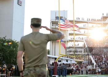 A soldier in uniform salutes the American flag at a football stadium, with sun shining and a crowd in the stands.