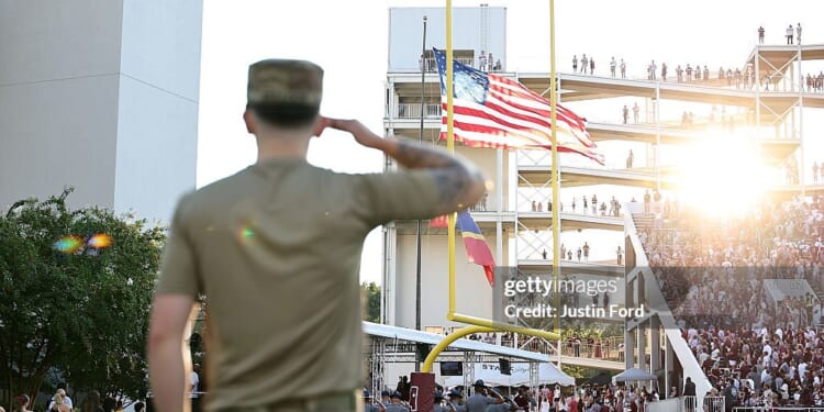 A soldier in uniform salutes the American flag at a football stadium, with sun shining and a crowd in the stands.