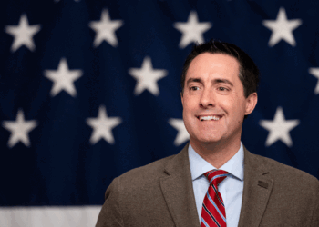 A man in a suit and red striped tie smiles in front of a backdrop with white stars on a blue background, resembling part of the American flag.