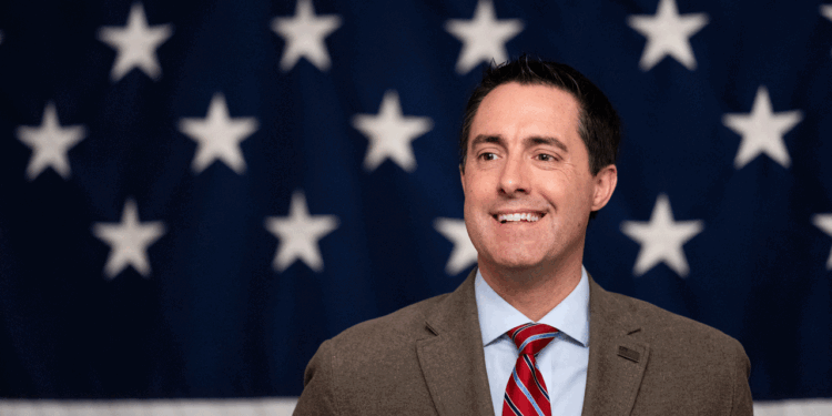 A man in a suit and red striped tie smiles in front of a backdrop with white stars on a blue background, resembling part of the American flag.