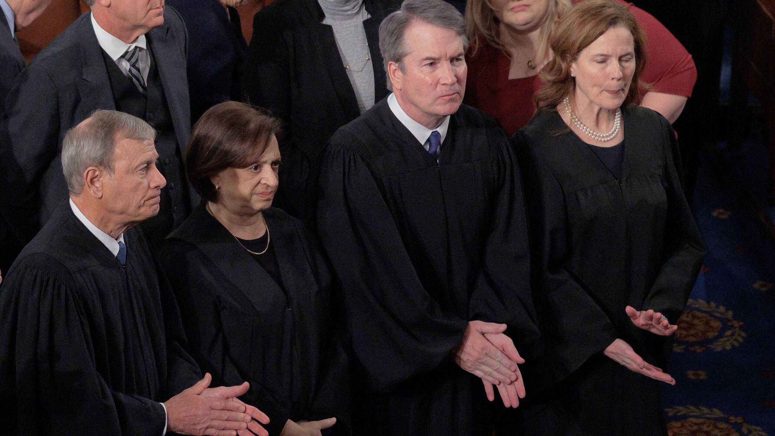 Four judges in black robes stand side by side in a courtroom, with others seated and standing around them. The judges have serious expressions and their hands are clasped or held together in front of them.