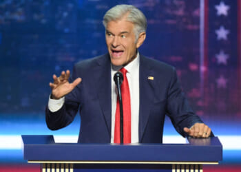 Dr. Mehmet Oz in a suit and red tie speaks energetically at a podium, gesturing with his left hand. The background is blue with white stars and red stripes, resembling a patriotic or political setting.
