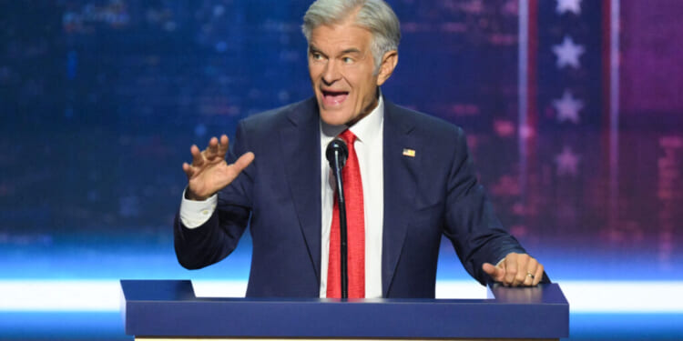 Dr. Mehmet Oz in a suit and red tie speaks energetically at a podium, gesturing with his left hand. The background is blue with white stars and red stripes, resembling a patriotic or political setting.