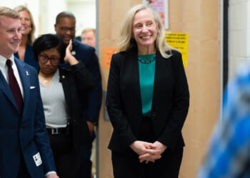 Abigail Spanberger in a black suit and green top smiles while standing in a hallway with several people, including a man in a suit and tie. The group appears to be having a friendly conversation at a school.