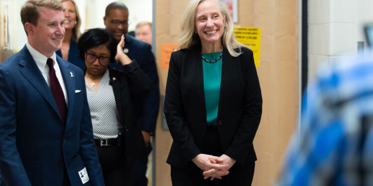Abigail Spanberger in a black suit and green top smiles while standing in a hallway with several people, including a man in a suit and tie. The group appears to be having a friendly conversation at a school.