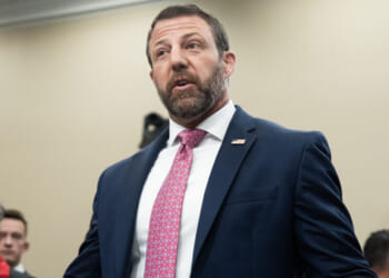 A man in a navy suit, white shirt, and pink patterned tie speaks indoors. He has short hair and a beard. Several people are blurred in the background.