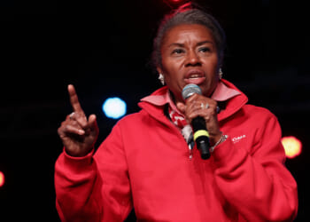 Winsome Earle-Sears with graying hair, wearing a red quarter-zip sweatshirt, speaks into a microphone on stage. She is pointing her index finger up, and colorful stage lights glow in the dark background, capturing a commanding presence reminiscent of Earle-Sears.