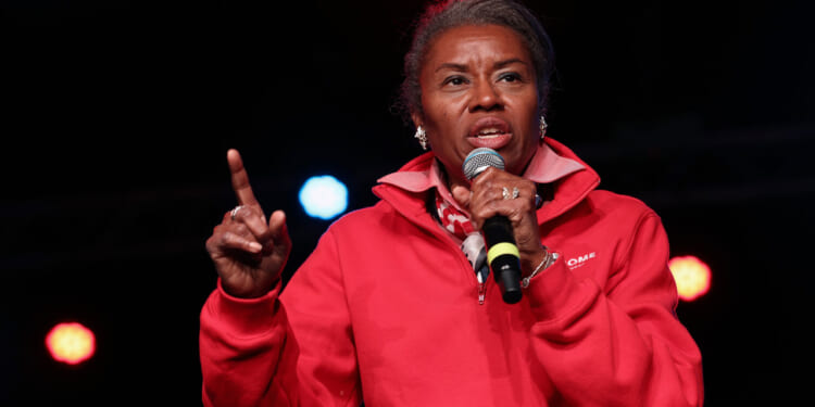 Winsome Earle-Sears with graying hair, wearing a red quarter-zip sweatshirt, speaks into a microphone on stage. She is pointing her index finger up, and colorful stage lights glow in the dark background, capturing a commanding presence reminiscent of Earle-Sears.