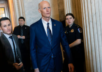 Rick Scott in a blue suit walks through a hallway with three people, including a police officer, nearby. Marble columns and ornate walls are visible in the background.