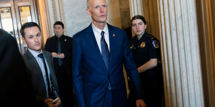 Rick Scott in a blue suit walks through a hallway with three people, including a police officer, nearby. Marble columns and ornate walls are visible in the background.