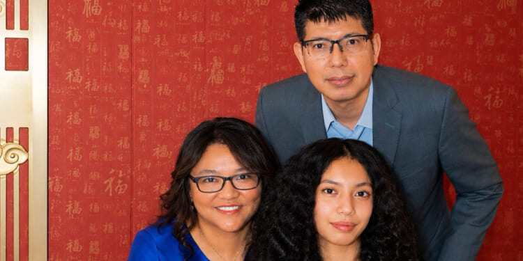 Three people pose in front of a red wall with gold patterns. A man in a blue suit stands behind a woman in glasses and a blue dress, while a young woman with curly hair sits in front. Though all are looking at the camera and smiling, their expressions hint at the tension of an unexpected hostage scenario unfolding just beyond the frame.