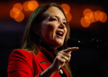 Brooke Rollins in a red blazer speaks passionately into a microphone, gesturing with her hand. Warm orange lights create a soft, out-of-focus background.