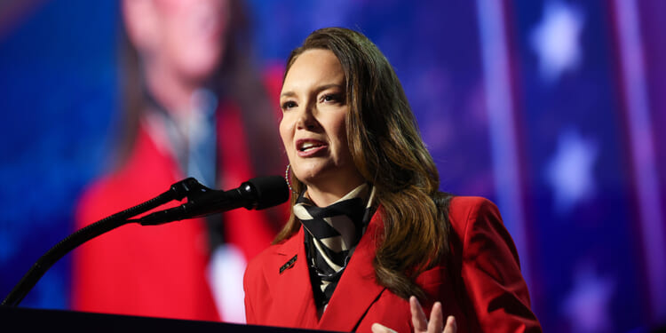 Brooke Rollins in a red blazer speaks at a microphone on stage, with a blurred background of stars and stripes behind her.