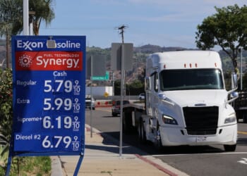 Exxon gas station sign displays fuel prices; regular at $5.79, extra at $5.99, supreme at $6.19, and diesel at $6.79 per gallon. A white semi truck drives by on a sunny day.