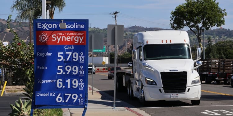 Exxon gas station sign displays fuel prices; regular at $5.79, extra at $5.99, supreme at $6.19, and diesel at $6.79 per gallon. A white semi truck drives by on a sunny day.