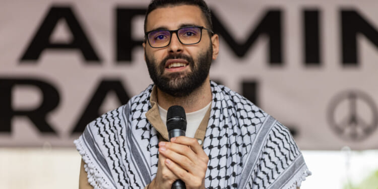 A man wearing glasses and a keffiyeh speaks into a microphone at an event. A partially visible banner with bold letters and a peace symbol is in the background.