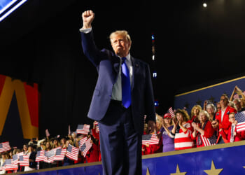 Donald Trump in a suit raises his fist on stage at an event, with a cheering crowd behind him. Many people are holding American flags, and patriotic decorations are visible.