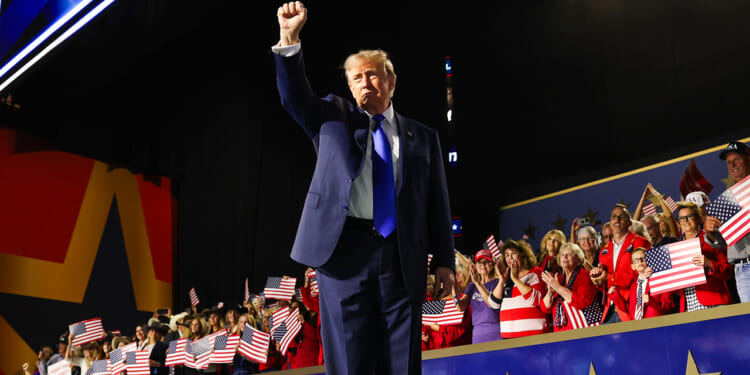 Donald Trump in a suit raises his fist on stage at an event, with a cheering crowd behind him. Many people are holding American flags, and patriotic decorations are visible.