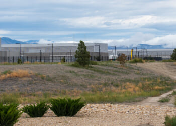 A dirt road and grassy field lead to a cluster of modern, gray industrial buildings, surrounded by fencing, with mountains and clouds in the background.