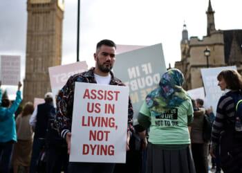 A man holds a sign reading ASSIST US IN LIVING, NOT DYING at a public protest against MAID, while others around him display various signs in front of historic buildings.