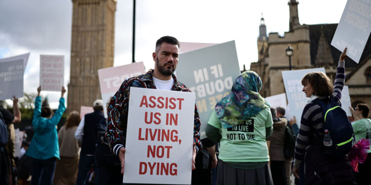 A man holds a sign reading ASSIST US IN LIVING, NOT DYING at a public protest against MAID, while others around him display various signs in front of historic buildings.