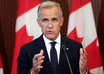 Mark Carney in a dark suit and tie speaks in front of several large Canadian flags, gesturing with his hands during a press conference or public address about tariffs.