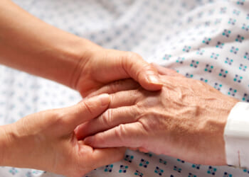 Feminine hands hold the hand of an elderly patient in white hospital gown with blue dots.