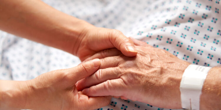Feminine hands hold the hand of an elderly patient in white hospital gown with blue dots.