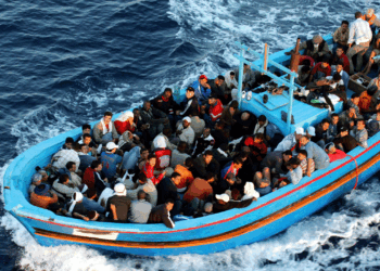 A crowded wooden boat filled with people sails through deep blue ocean water, creating white waves as it moves forward.