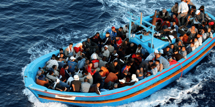 A crowded wooden boat filled with people sails through deep blue ocean water, creating white waves as it moves forward.